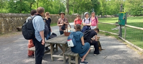 Eine Gruppe von Menschen steht und sitzt um einen Holztisch herum, ein Mann mit einem grünen Hut in der Mitte der Gruppe erzählt etwas.