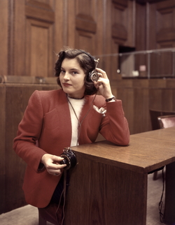 Eine junge Frau mit einer roten Jacke sitzt an einem Holztisch, dreht mit der rechten Hand an einem Drehknopf, der seitlich am Tisch befestigt ist, und hält mit der linken ihre Kopfhörer